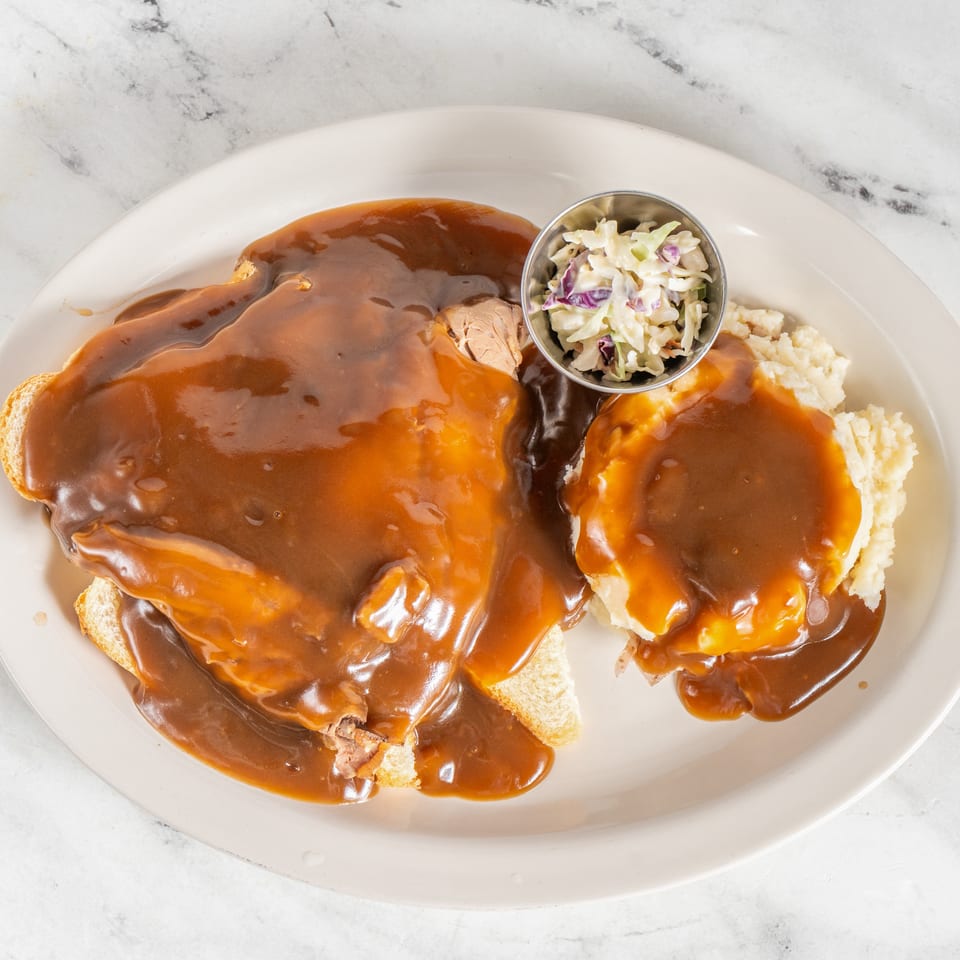 Open-faced roast beef sandwich and mashed potatoes covered in brown gravy, served with creamy coleslaw on a white plate.