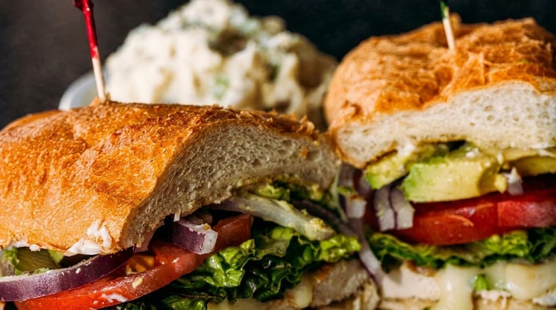 Two halves of a chicken sandwich with lettuce, tomato, onion, avocado, and cheese, with potato salad in the background.