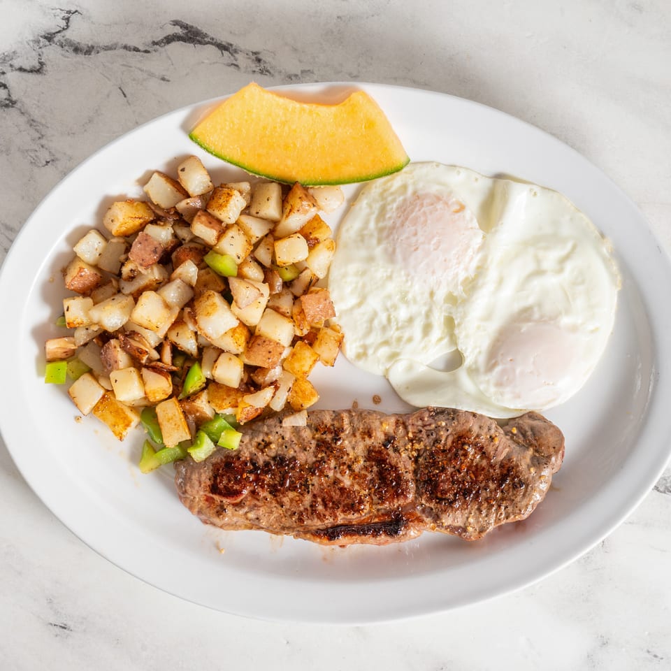 Grilled steak served with fried eggs, diced potatoes and peppers, and a slice of cantaloupe on a white plate.