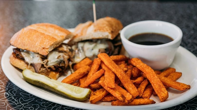 Philly cheesesteak sandwich with melted cheese, sweet potato fries, a pickle spear, and au jus.
