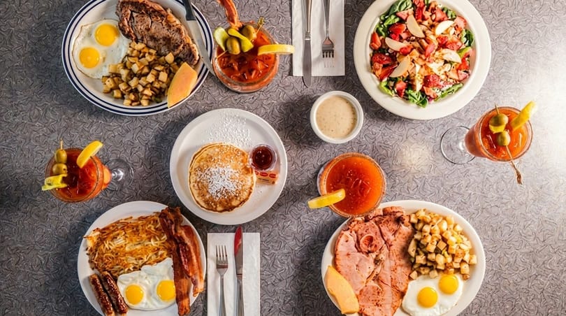 Overhead view of various breakfast dishes including eggs, bacon, pancakes, steak, and Bloody Marys.