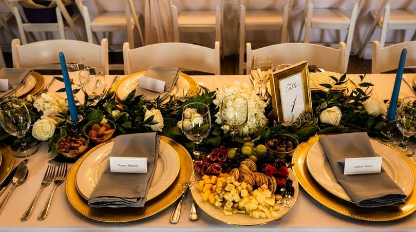 Elegantly set dining table with gold chargers, floral garland, blue candles, and a charcuterie board.