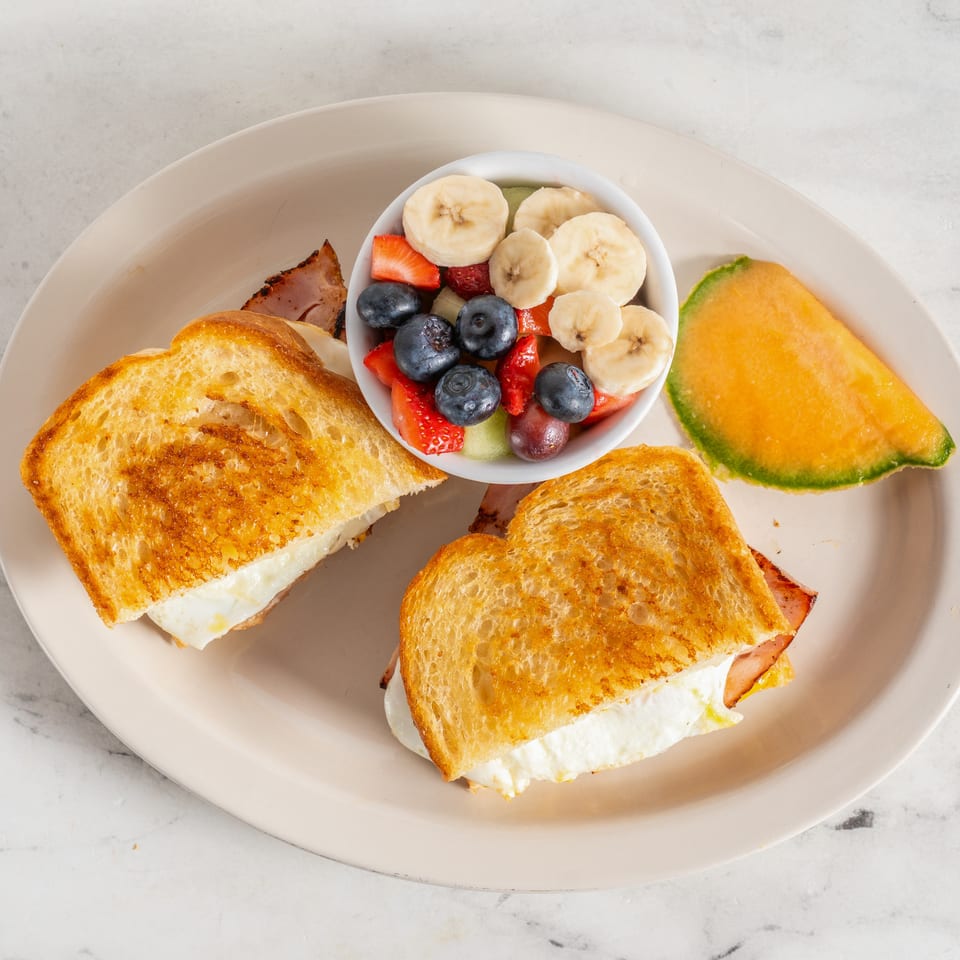 Breakfast plate with toasted sandwiches, mixed fruit, cantaloupe, and bacon on a pale dish.