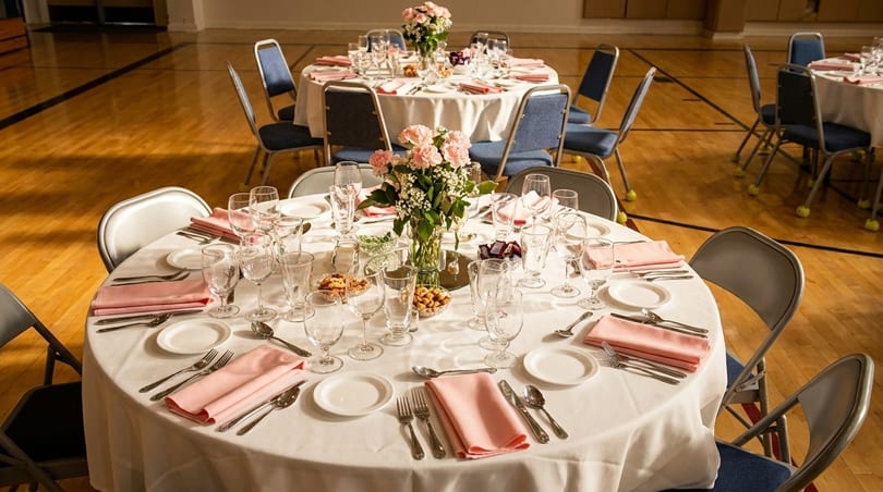 Round tables set with white tablecloths, pink napkins, and floral centerpieces in a gymnasium.