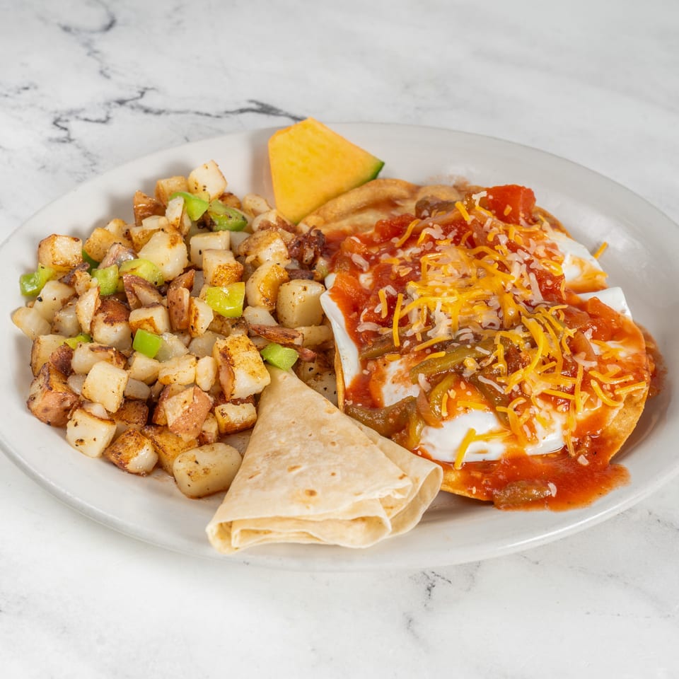 Breakfast plate with seasoned home fries, green peppers, tortillas, cantaloupe, and a cheesy salsa-topped egg dish.