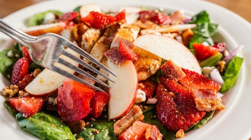 A fork in a vibrant salad with strawberries, apples, chicken, bacon, and spinach.
