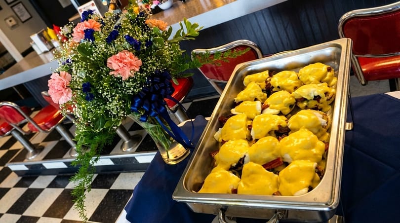A buffet tray of eggs benedict with hollandaise sauce, next to a floral arrangement in a diner setting.