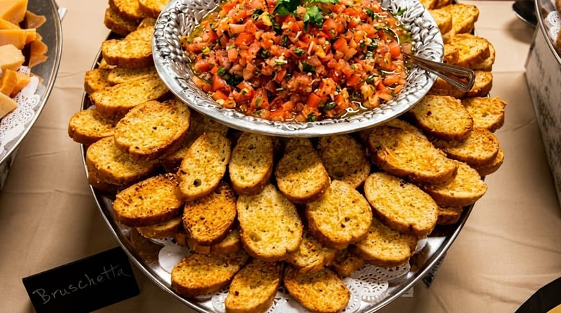 A silver platter of golden bruschetta toasts surrounds a bowl of fresh tomato bruschetta topping.