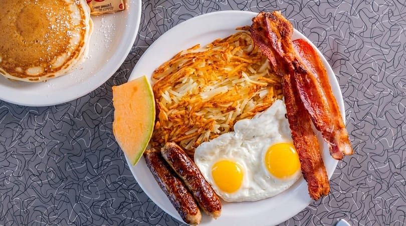 A full breakfast plate with hash browns, bacon, sausage, fried eggs, and a side of melon.