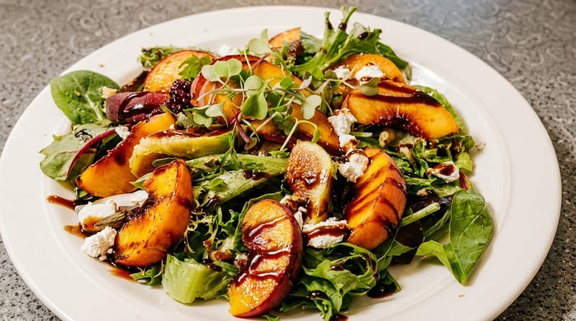 Fresh salad with peaches, figs, goat cheese, mixed greens, and balsamic glaze on a white plate.